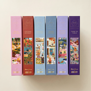 Six boxed jigsaw puzzle books are lined up against a warm off-white background. The spines are in different pastel and earthy colors: pink, terracotta, light blue, medium blue, lavender, and deep purple.