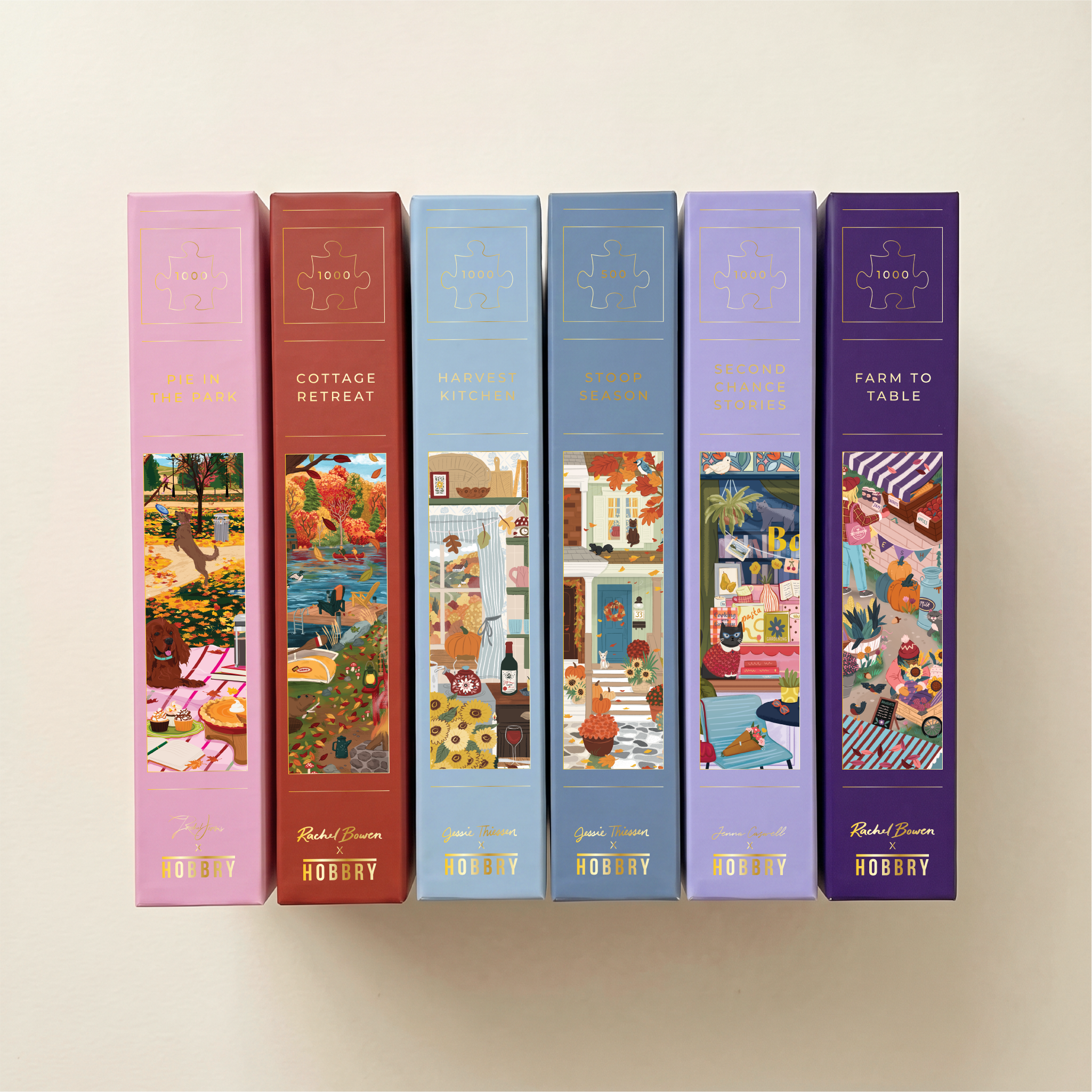 Six boxed jigsaw puzzle books are lined up against a warm off-white background. The spines are in different pastel and earthy colors: pink, terracotta, light blue, medium blue, lavender, and deep purple.