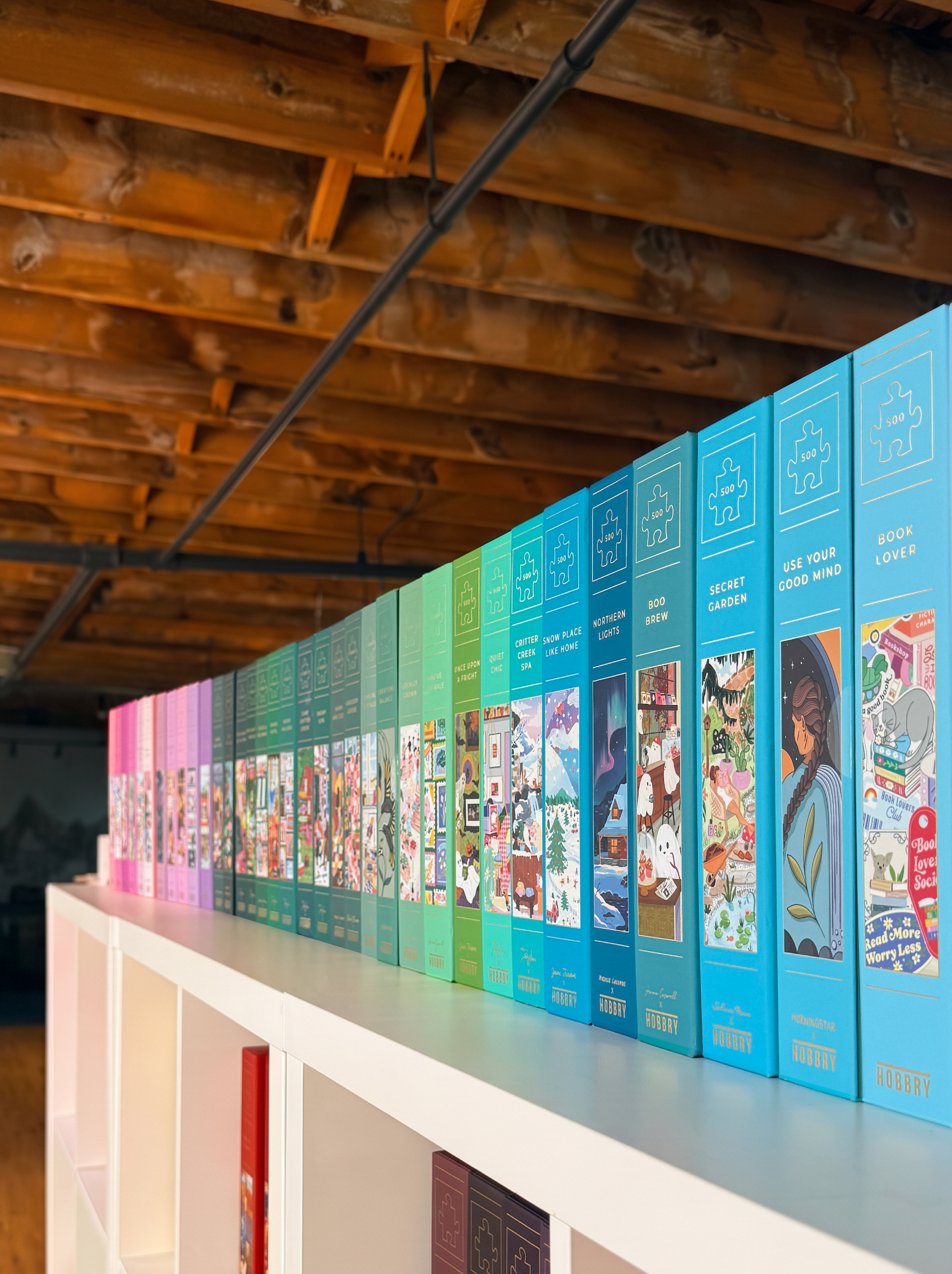 A row of colorful jigsaw puzzle boxes arranged on a white shelf in an indoor space with exposed wooden ceiling beams. The boxes are in various bright colors including blue, green, teal, and pink, with puzzle titles and illustrations visible on the front.
